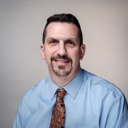 Headshot of Keith Gnoza looking directly at the camera while wearing a blue shirt and brown paisley tie.