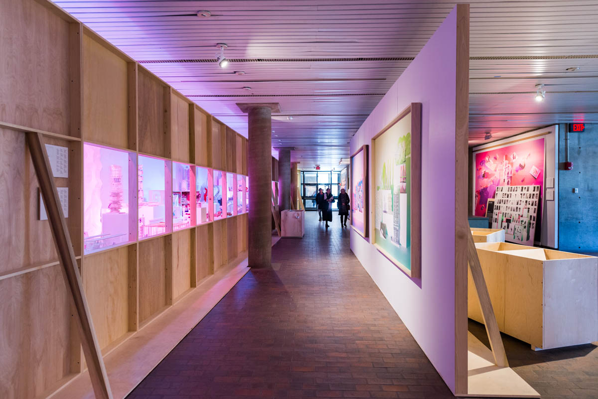 A view down a constructed hallway. One wall is a plain plywood windowed wall showcasing select student models cross-it in blue and pink hues, and the other has two hanging photos.