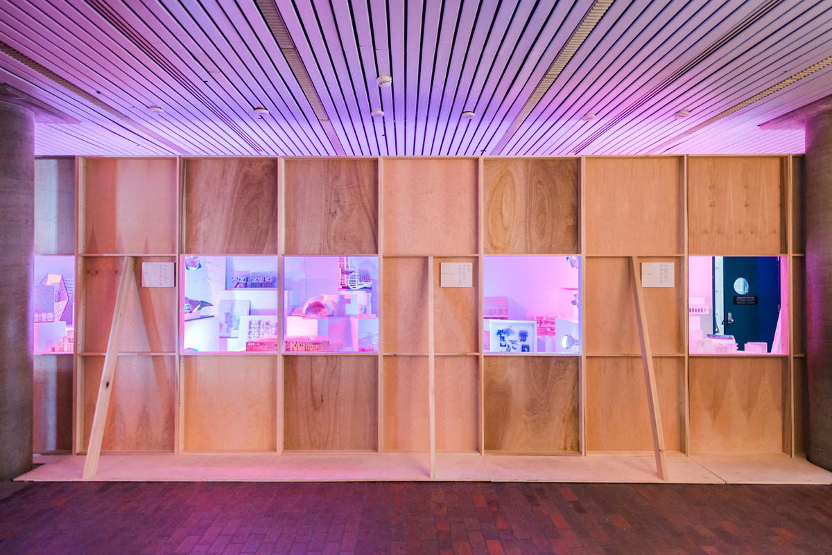 A plain plywood wall obstructing student models brightly cross lit in blue and pink hues. Square windows showcase selected views.