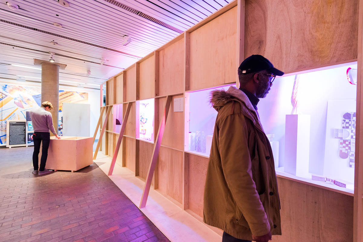 A person looking through the square windows of a plain plywood wall obstructing student models brightly cross lit in blue and pink hues. Another person on is looking through flat student work stacked in a plywood crate.