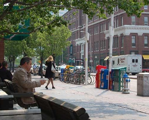 People stand and sit on benches along the sidewalk of a street.