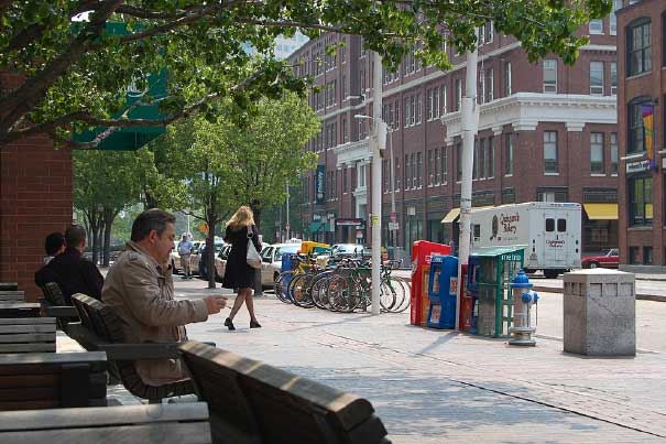 People stand and sit on benches along the sidewalk of a street.