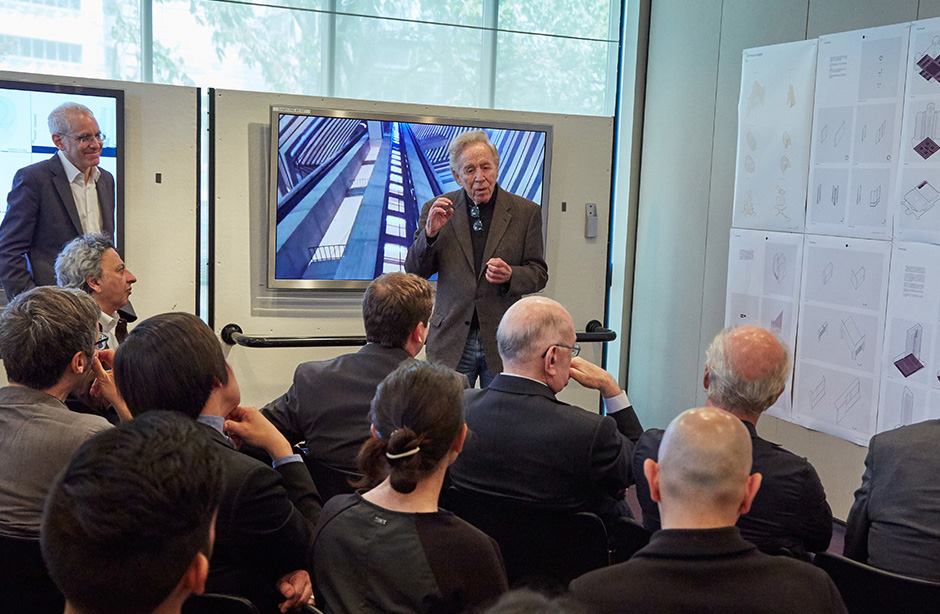 Man speaking to audience in front of pictures elf hotel atrium with visible elevators.