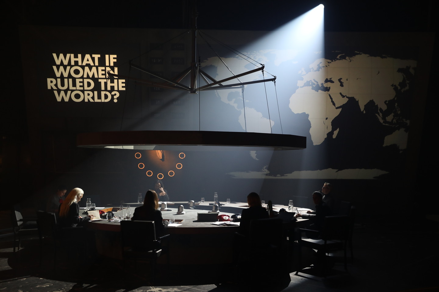 An image of women seated around a circular table. The text "what if women ruled the world?" is above them.