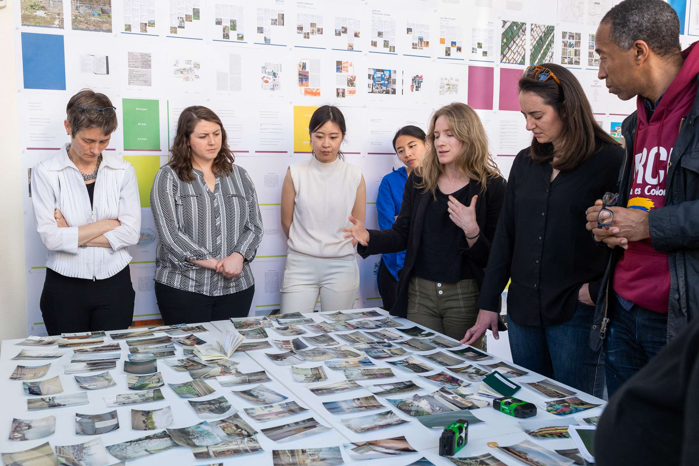 7 people stand around a square table covered with dozens of small color photographs.