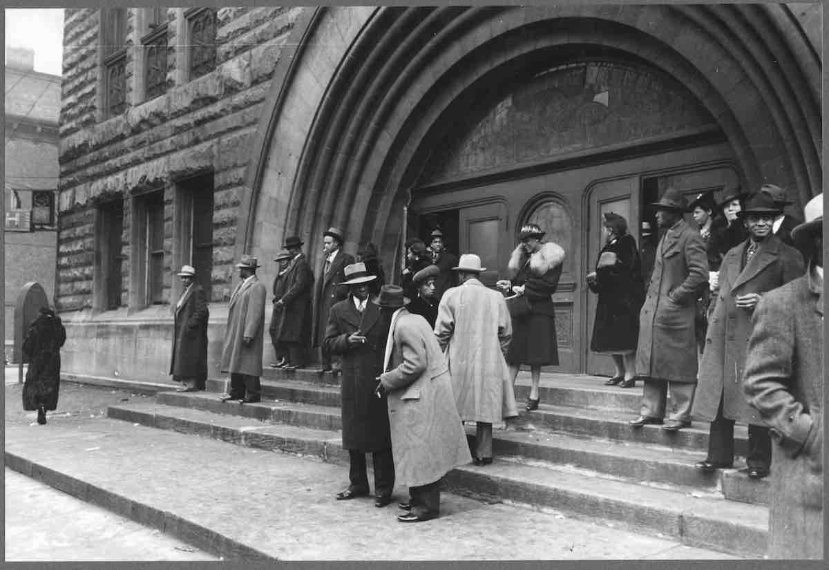 A black and white image of a group of people in long coats exiting a church, underneath a large archway.