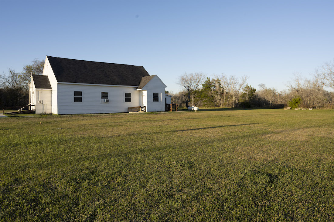 A white church with a gray roof sits in the middle left of the image on a large, open grassy area, with sparse trees in the background.