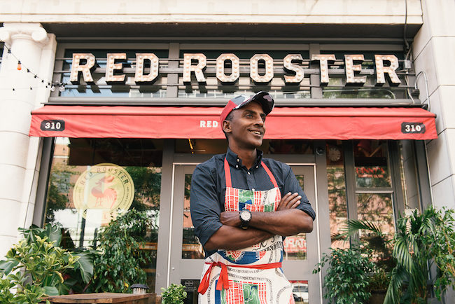 Marcus Samuelsson stands outside with his arms crossed, in front of his restaurant Red Rooster. The restaurant sign is above his head, with plants on either side of him. He wears a baseball cap, a blue-gray collared shirt, and a colorful apron.