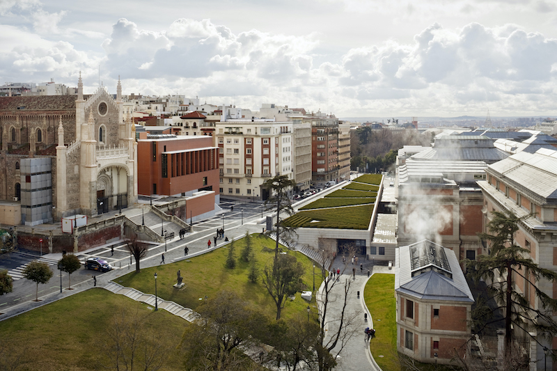 A landscape view of a city, with a road and green area running through the middle, fluffy clouds in the sky, and large buildings.