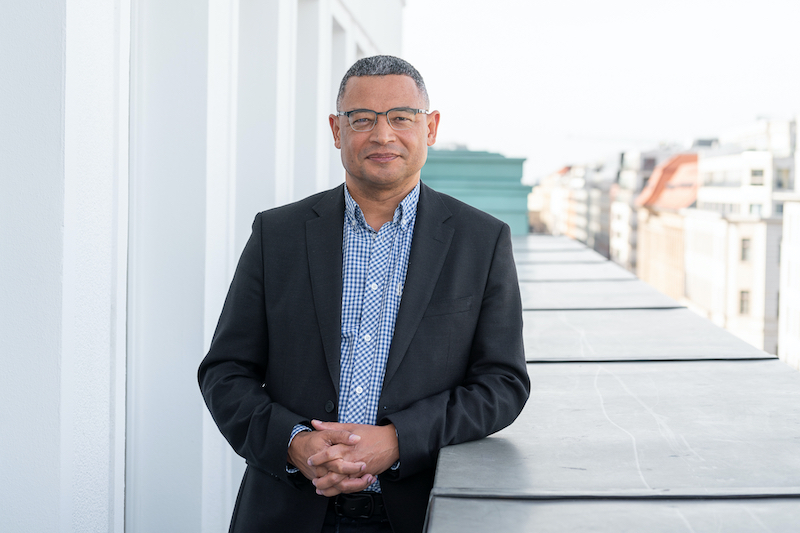 Edgar Pieterse, wearing glasses, a blue shirt, and a black blazer, stands with is hands clasped together, leaning on a ledge on the balcony of a large building.