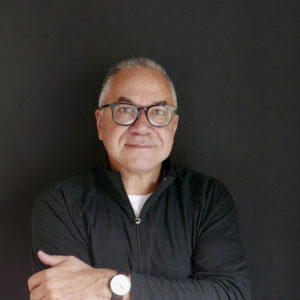 A photograph of a man with short white hair wearing glasses, a black long sleeved half-zip shirt. He's crossing his arms in front of him and a watch with a large white face is visible. 