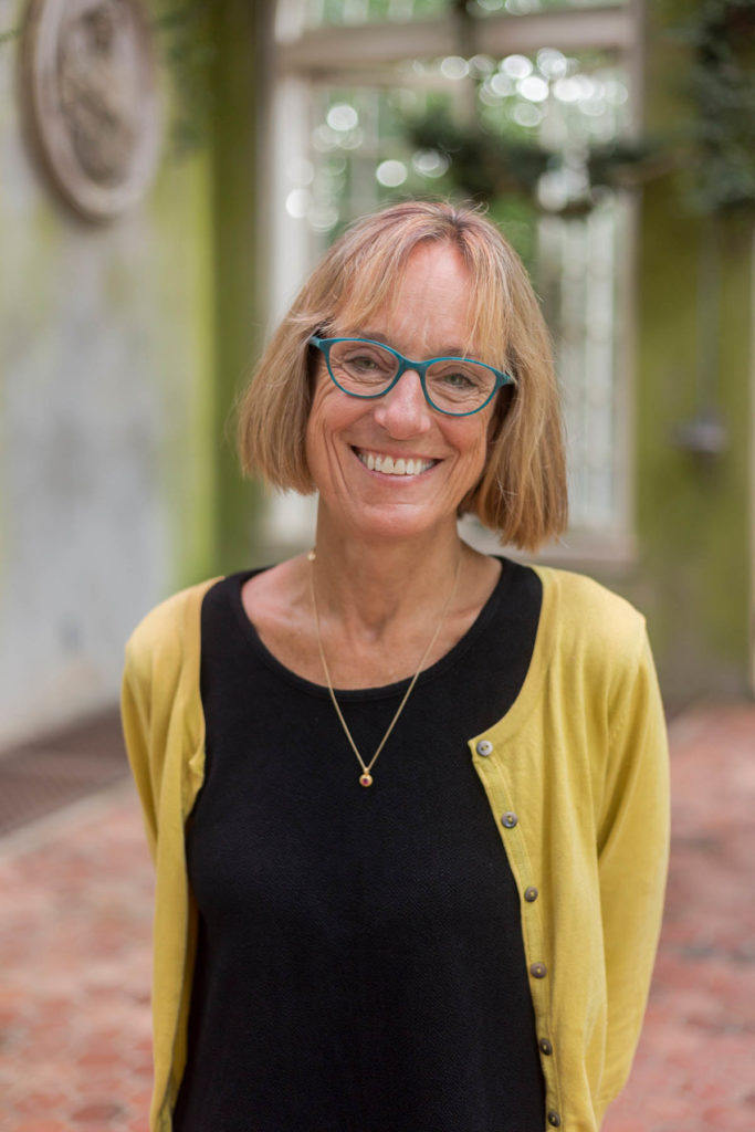 Headshot of Thaisa Way, who wears blue glasses, a black shirt, and a yellow sweater, and has chin-length blonde hair.