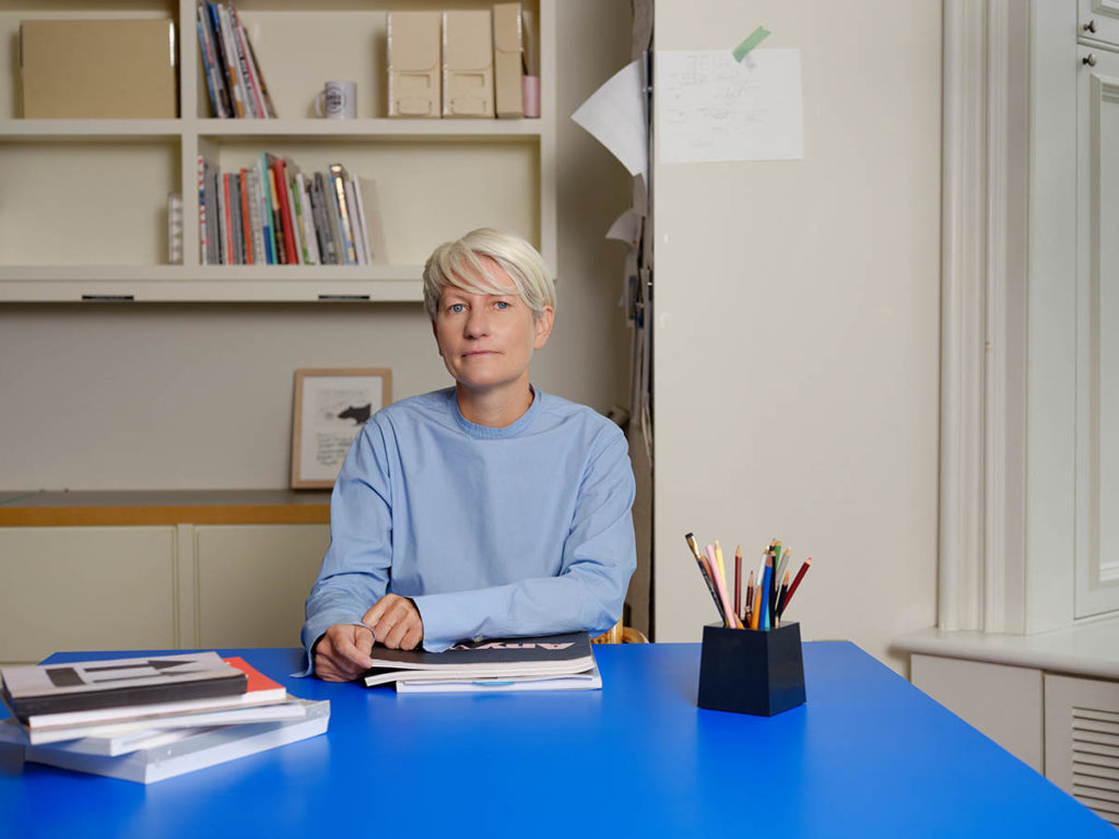 Image of Giovanna Borasi, who wears a light blue sweatshirt and sits at a dark blue desk. There are books stacked in front of her along with a variety of pens and pencils in a black square holder.