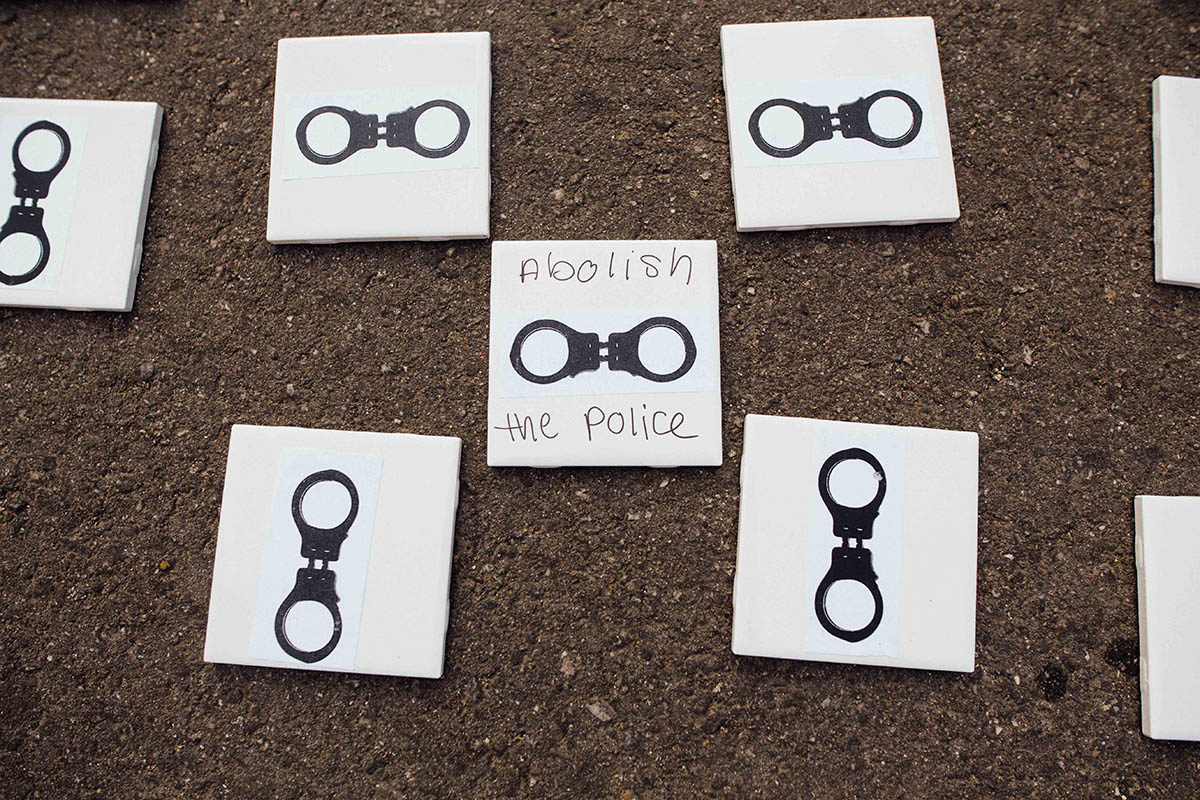 Photograph of five tiles laid on the ground. Each white tile has an image of handcuffs. On one of the tiles, someone has written 