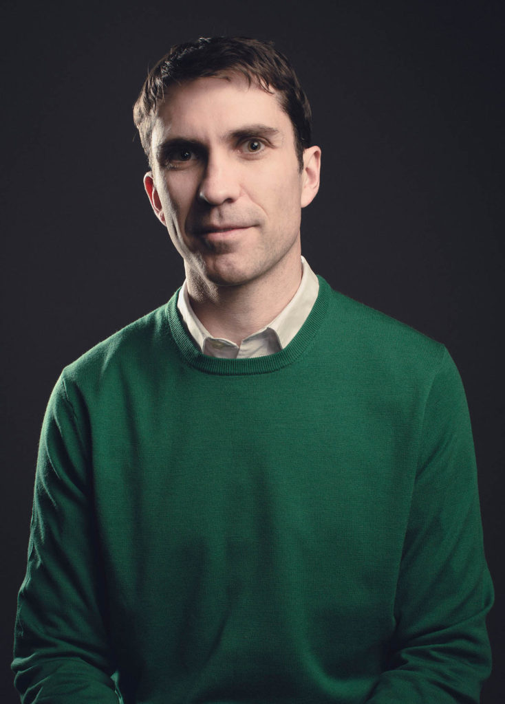 Headshot of Paul Anderson, who wears a green sweater over a white collared shirt and stands in front of a black dark gray background.