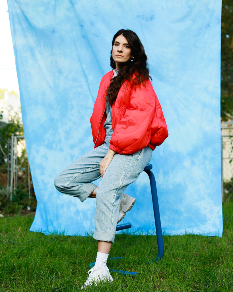 Portrait photo of Maite Borjabad Lopez-Pastor, who sits on a stool in front of a blue backdrop and wears a bright pink coat, grey pants, and white sneakers.