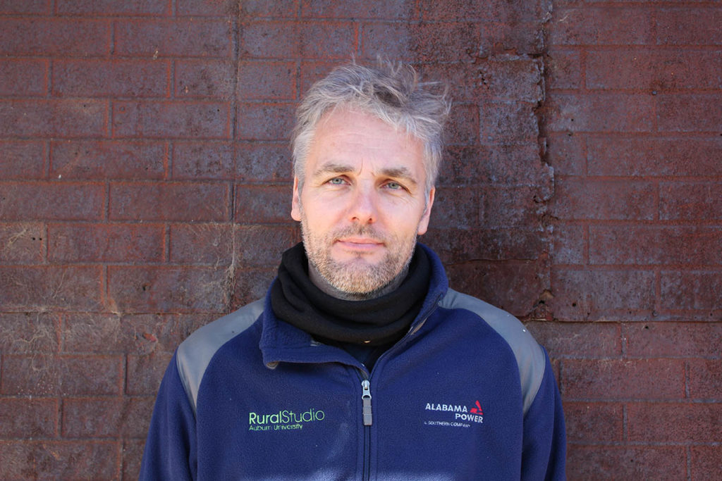 Headshot of Andrew Freear, who stands against a brick wall and wears a blue jacket.