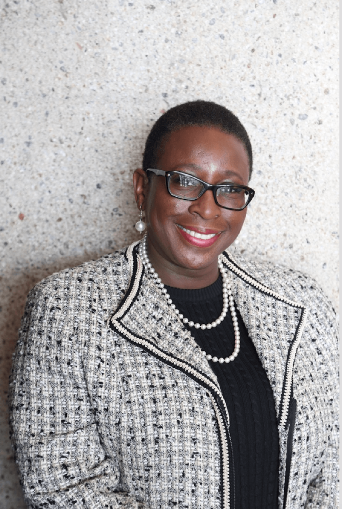 Headshot of Mayor Spicer, who wears black glasses, a gray checkered coat, a black shirt, and a pearl necklace.