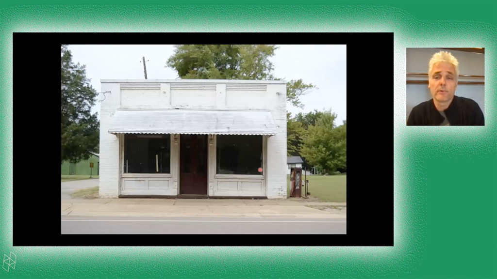 Screenshot from a virtual event. Andrew Freear appears in a small square on the right and wears a gray shirt and black jacket. A larger rectangle shows Andrew's PowerPoint presentation, which contains a photo of an old, small white building. Andrew and the PowerPoint are surrounded by a green background.