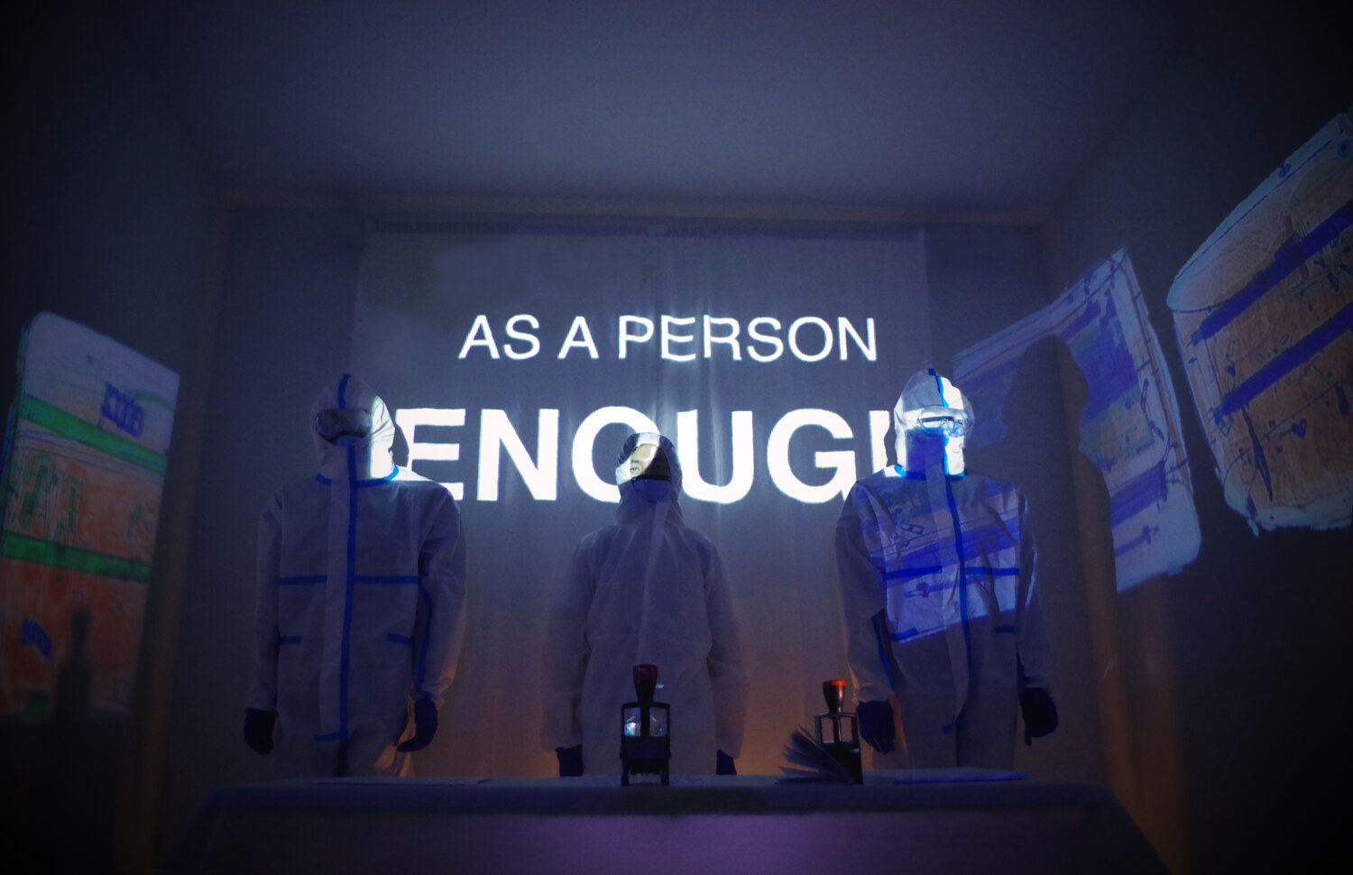 Three people wearing tyvek suits stand behind a table in a dark room lit by wall projections. On the table are two passport hand stamps. The words, "AS A PERSON ENOUGH" are projected onto the back wall.