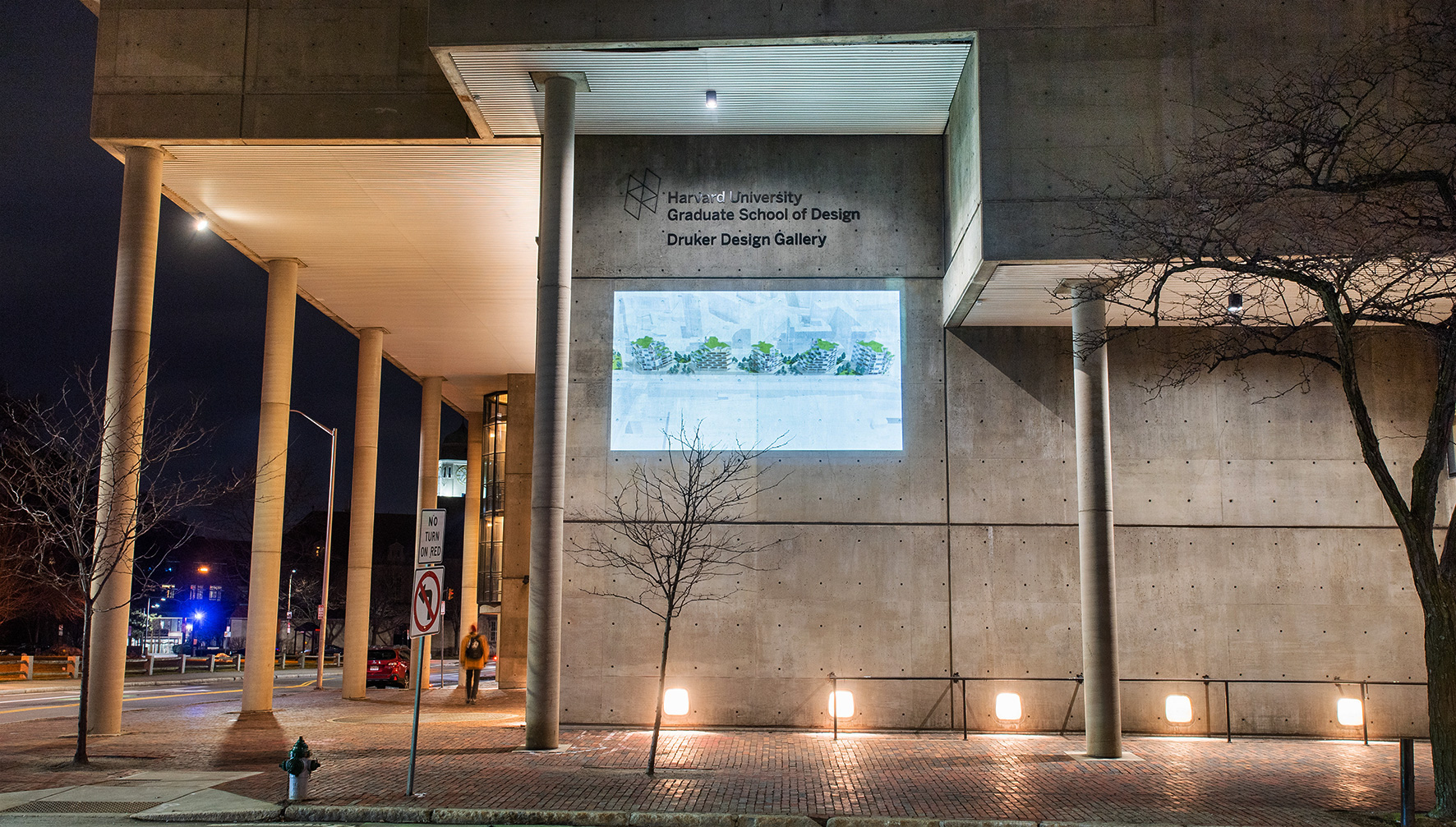The Cambridge Street facade of Gund hall at night. On the wall is projected an image of a birds-eye rendering of five Dual-Use buildings with balconies and grass-covered roofs.