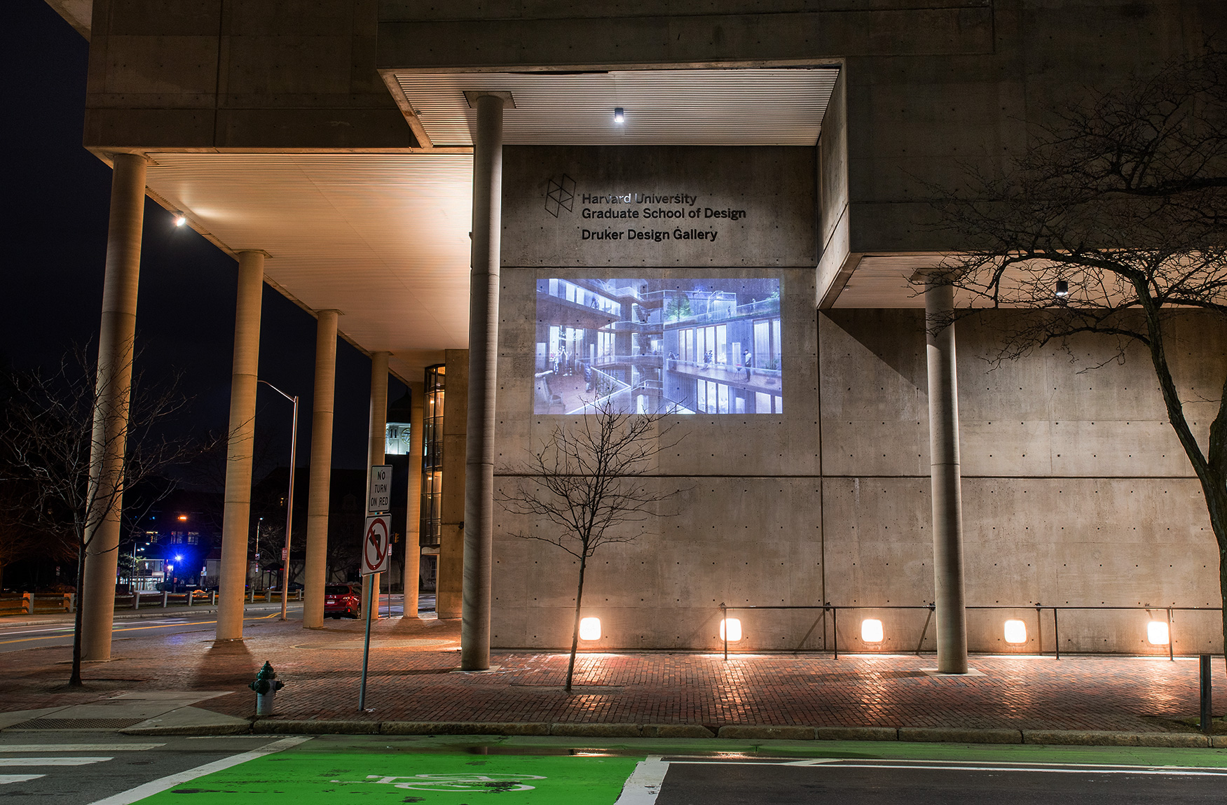 The Cambridge Street facade of Gund hall at night. On the wall is projected an image of a nighttime scene of the courtyard of a residential building with multiple levels of windows and balconies and people participating in various activities.