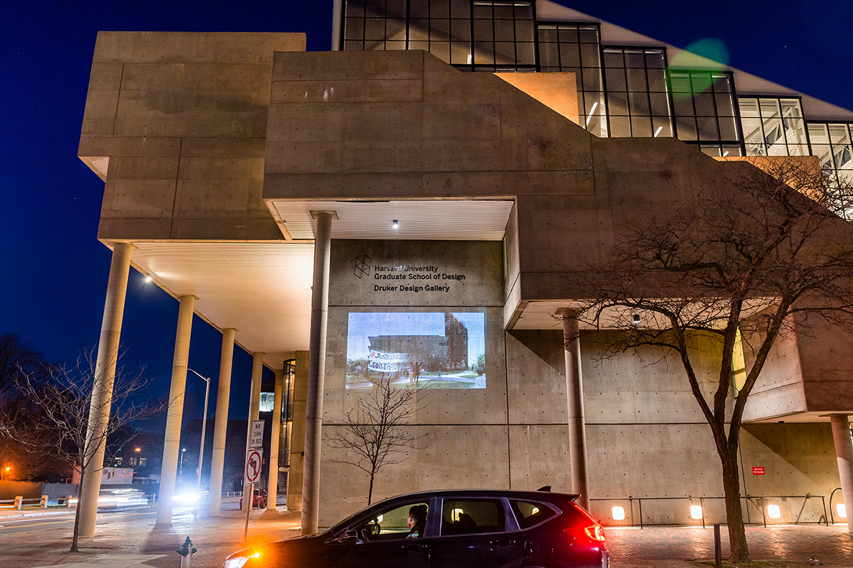 The Cambridge Street facade of Gund hall at night. On the wall is projected an image of a building with a demonstrator in front holding a sign that says “Justice for George Floyd”