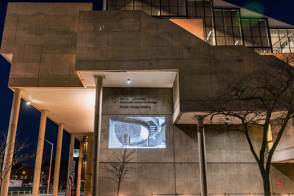 The Cambridge Street facade of Gund hall at night. On the wall is projected an image of a building facade with a curved stairwell coming down from an upper level.