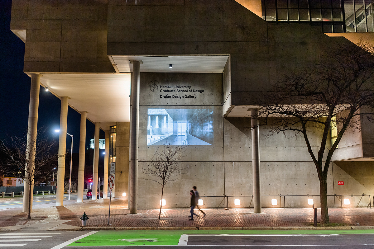 The Cambridge Street facade of Gund Hall at night, showing a projection of a person walking through an architectural interior space.
