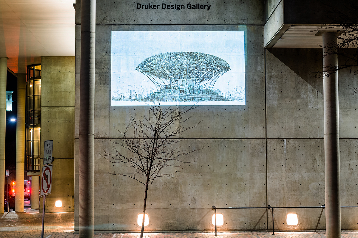The Cambridge Street facade of Gund Hall at night, showing a projection of a sketch drawing of a elliptical building on stilts.