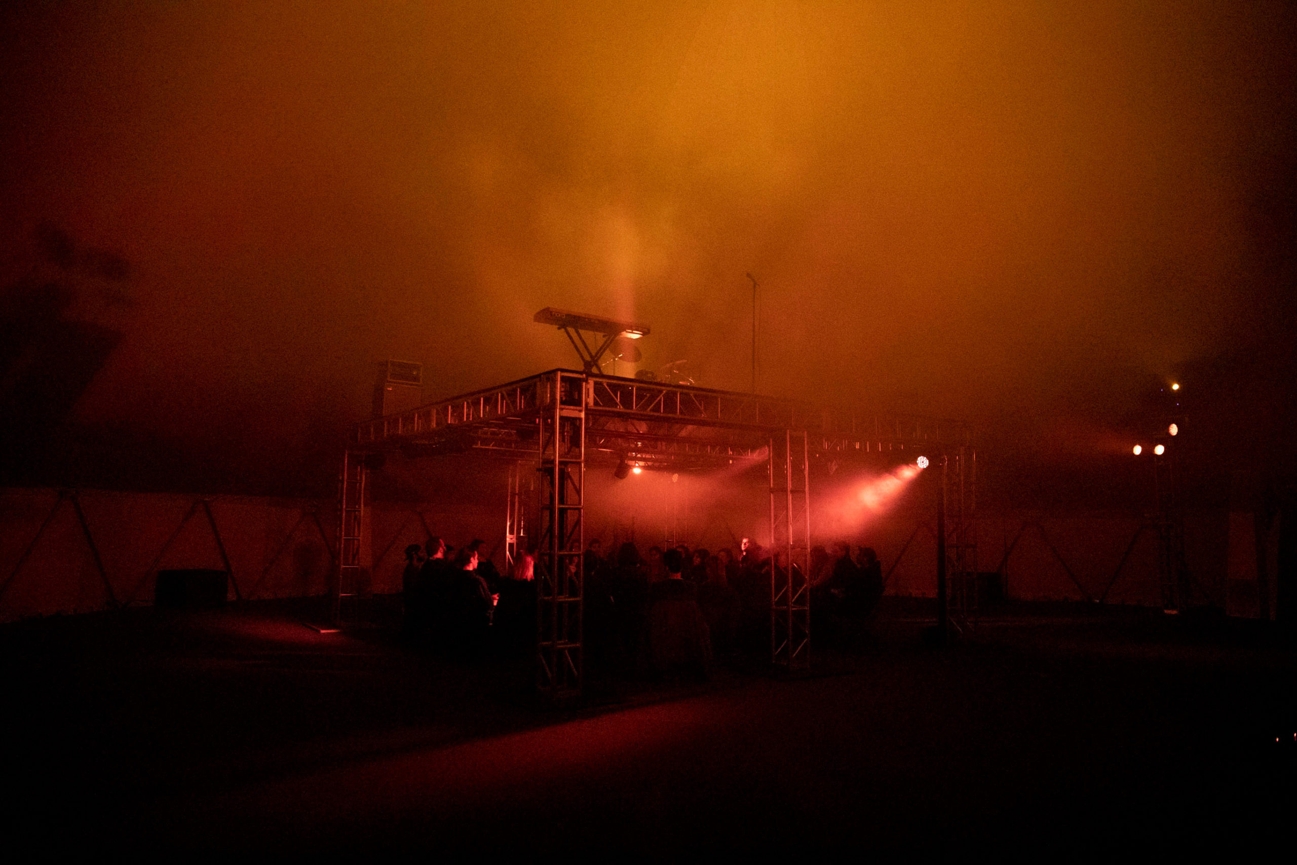 Image of interior scene of performance in a dark room with warm red and orange lights underneath a scaffold stage set up