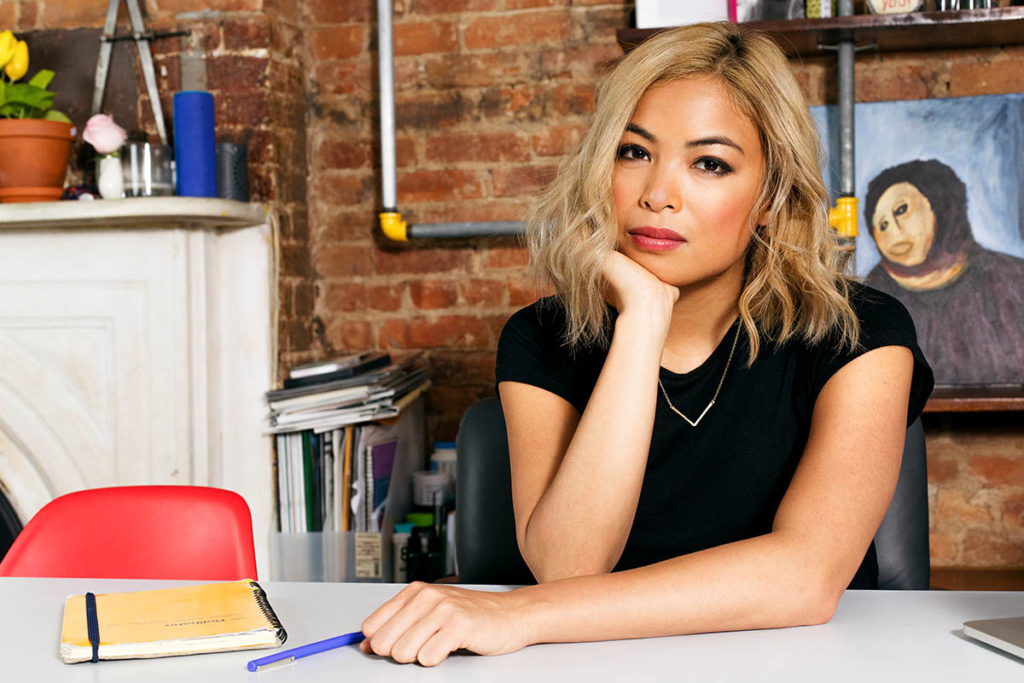 Headshot of Jia Tolentino, who wears a black shirt and has shoulder-length blonde hair. She sits at a desk with a pen and notebook, in front of a brick wall and mantle.