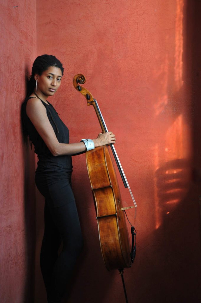 Photograph of Tomeka Reid, who wears all black and stands with a cello in front of a red-orange wall.