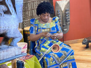 A seated woman wearing a blue and yellow patterned dress weaves a basket in a room containing African sculptures and textiles.