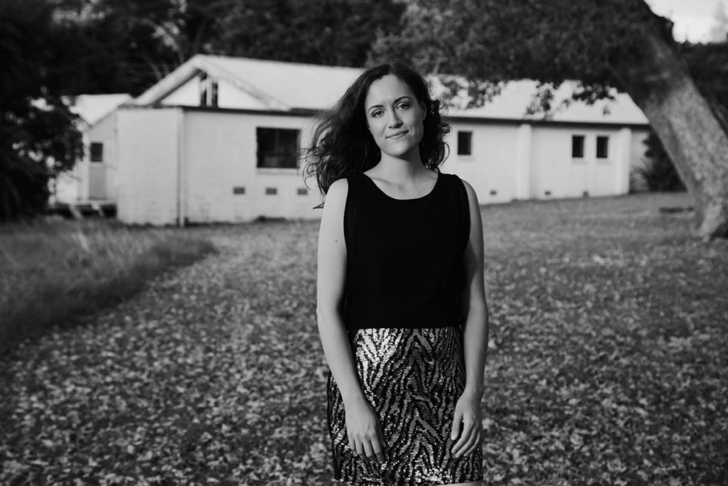 Black and white photograph of Jade Kake, who wears a black shirt and patterned skirt. She has long hair and stands in front of a white building, outdoors.