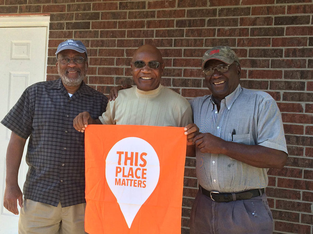 Image of three men standing in front of a brick wall holding a sign that says 