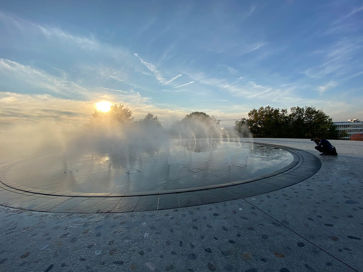 Photograph of a vast blue sky above a round water foundation, with someone crouched taking photos to the right.