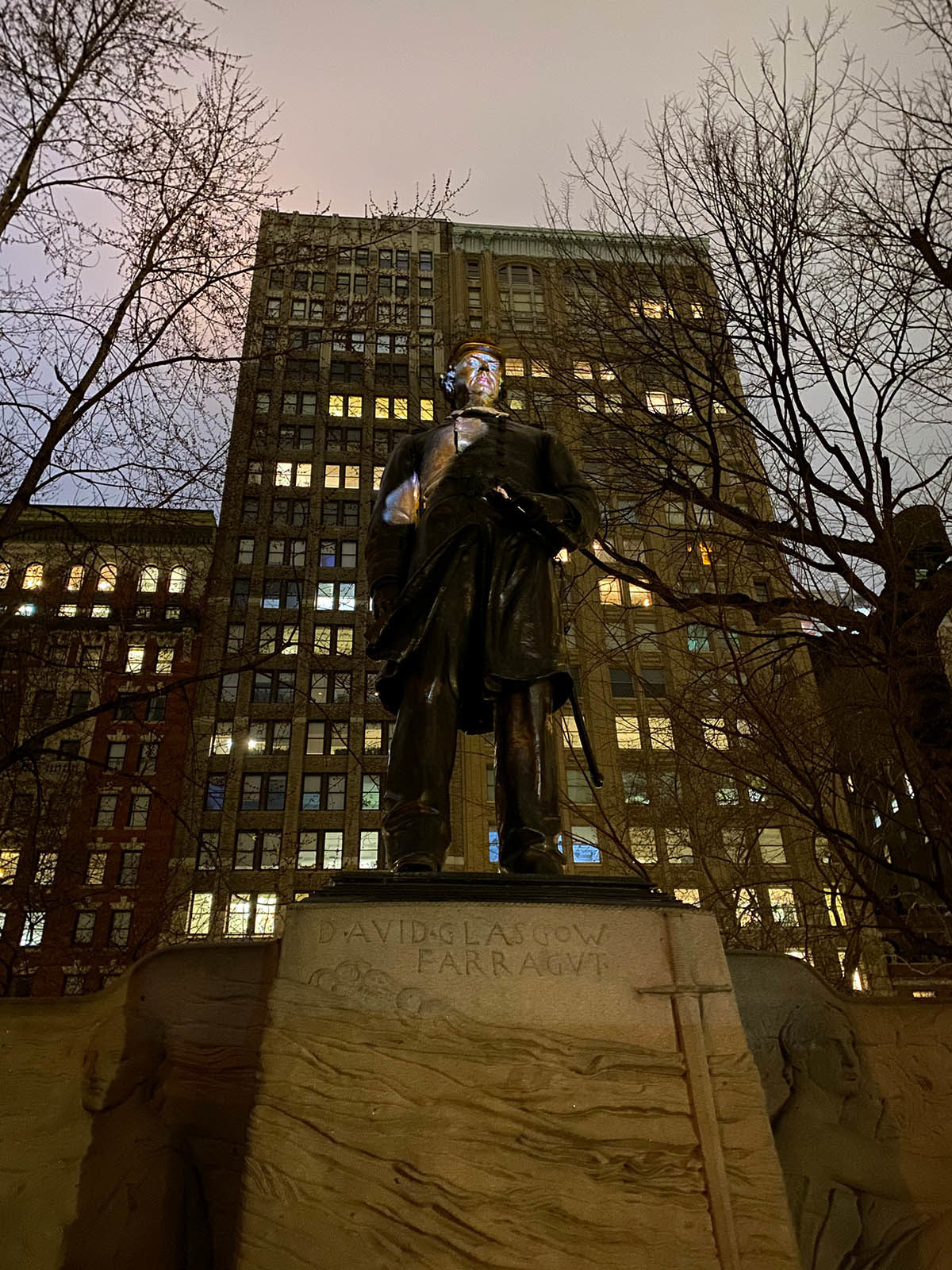 Photograph of a statue of a figure, seen from below. The statue stands in front of a tall building with many windows and a few sparse, neighboring trees.