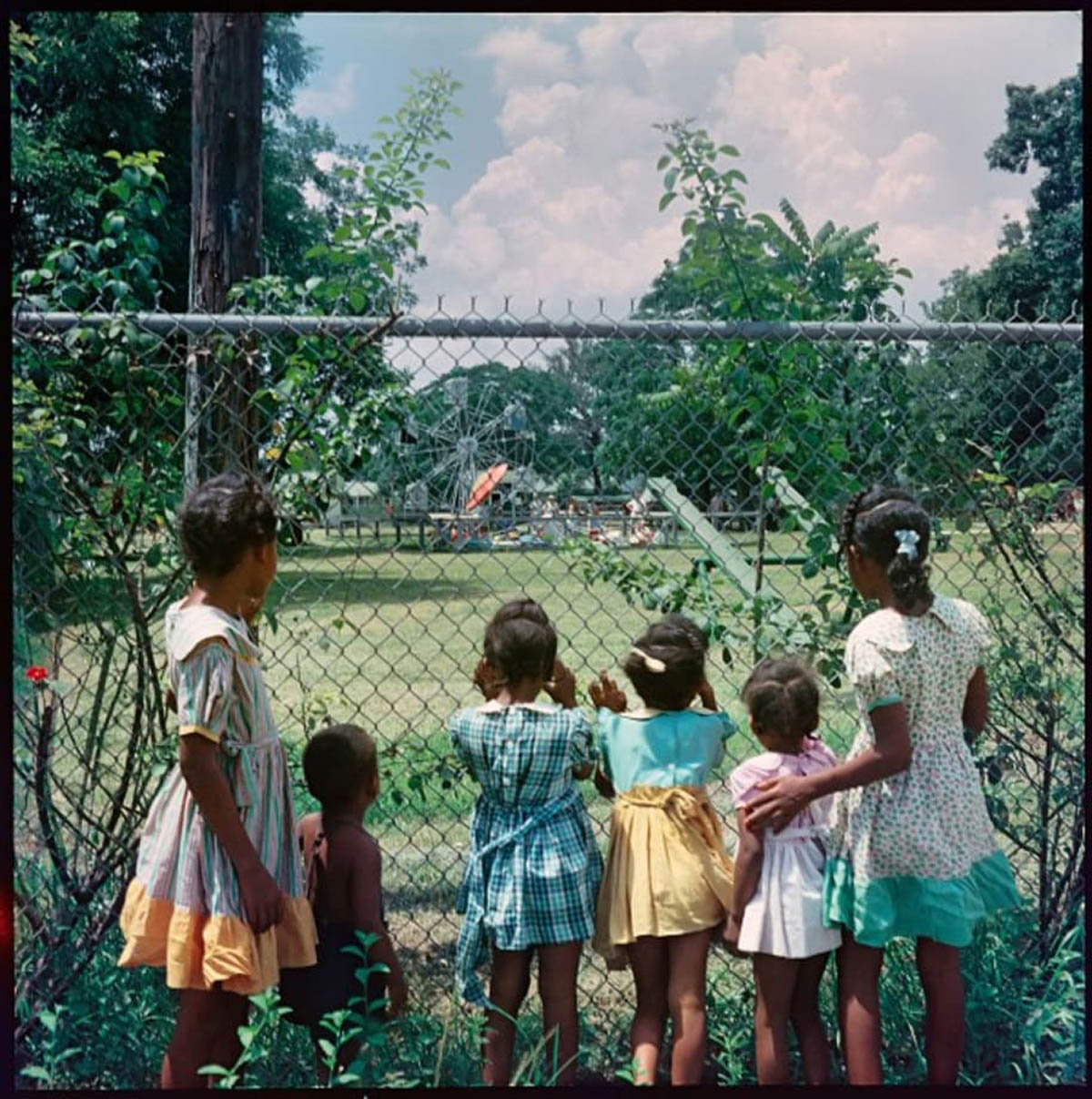 Photograph of six young children peering through a chainlink fence into a park.