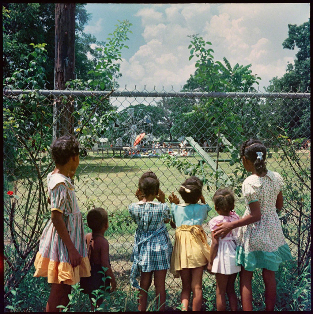 Photograph of six young children peering through a chainlink fence into a park.