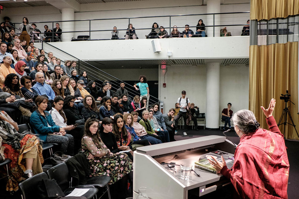 Photograph of a figure standing at a podium, from behind. The figure lectures to an audience of seated people.