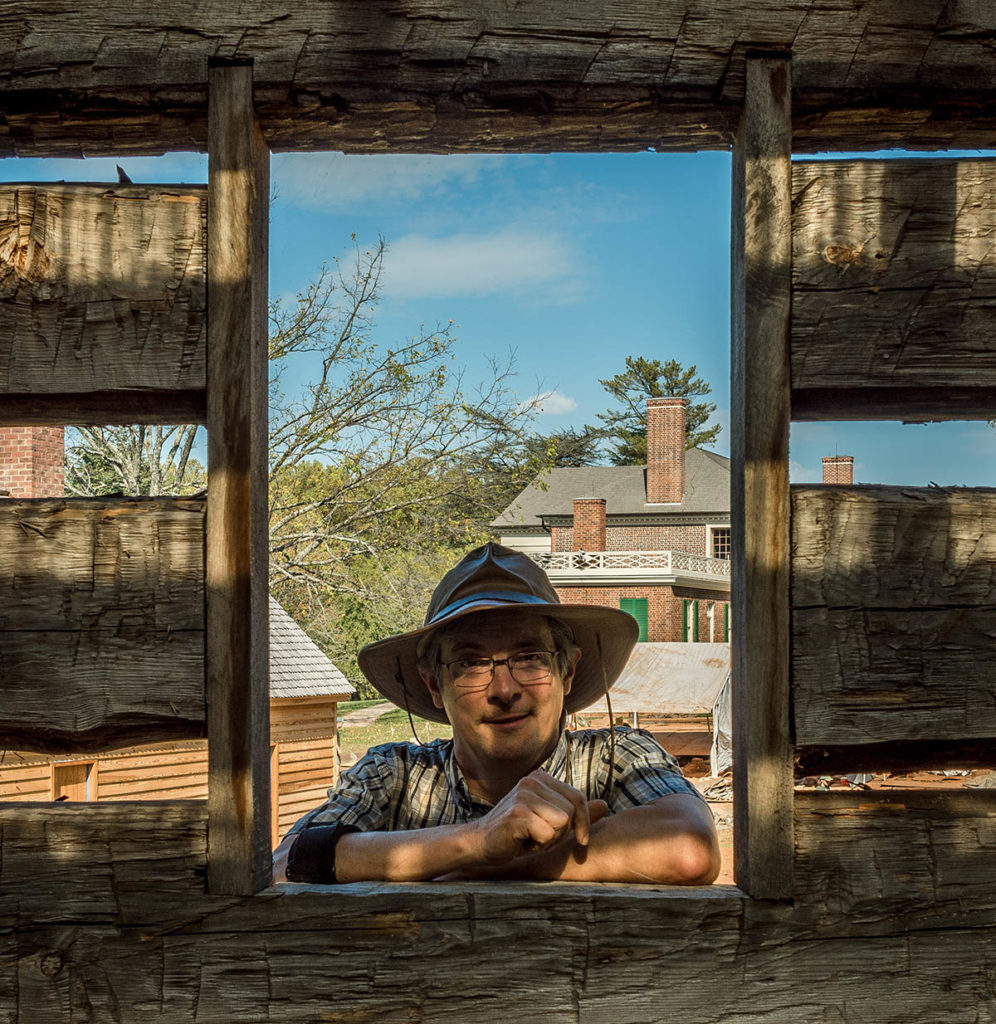 Photograph of Matthew Reeves, who looks through a window of a wood building and wears a hat and glasses.