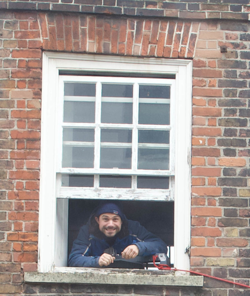 Photograph of Louis Schulz looking out a white window of a brick building.