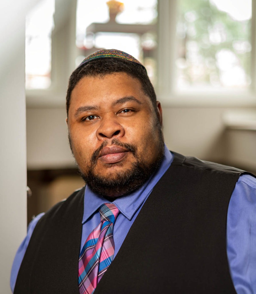 Headshot of Michael Twitty, who wears a black vest over a blue shirt with a pink and blue tie.