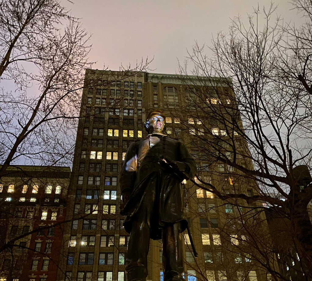 Photograph of a statue of a figure outdoors, in front of a tall building and surrounded by bare trees.