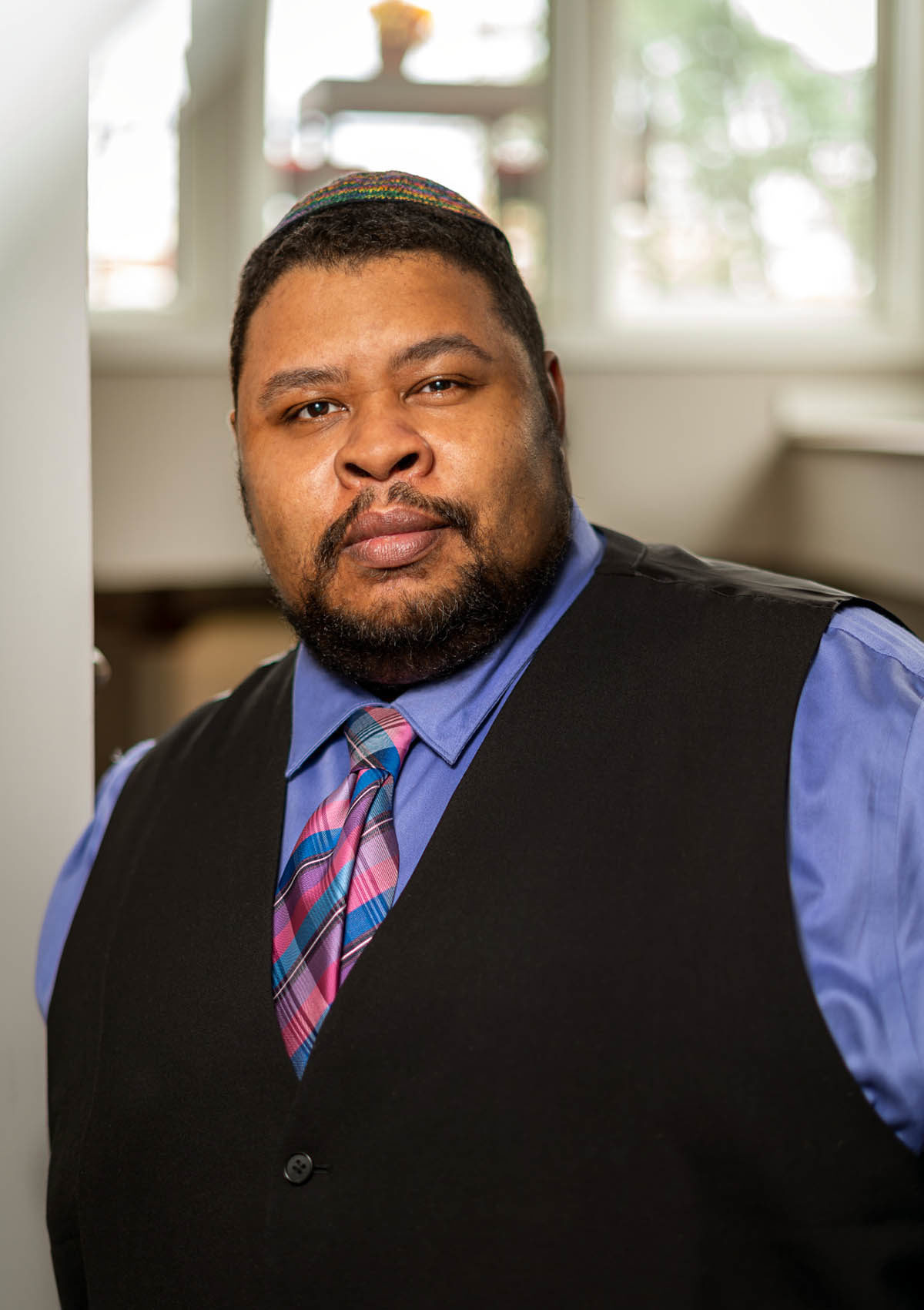 Headshot of Michael Twitty, who wears a black vest over a blue shirt with a pink and blue tie.