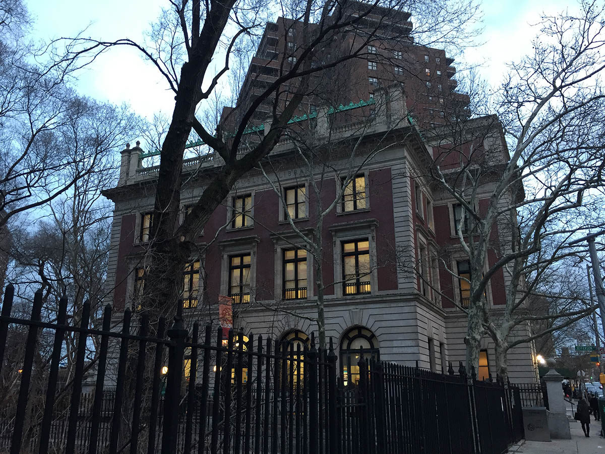 Photograph of a brick and stone building from the sidewalk, with a black gate and surrounding trees.