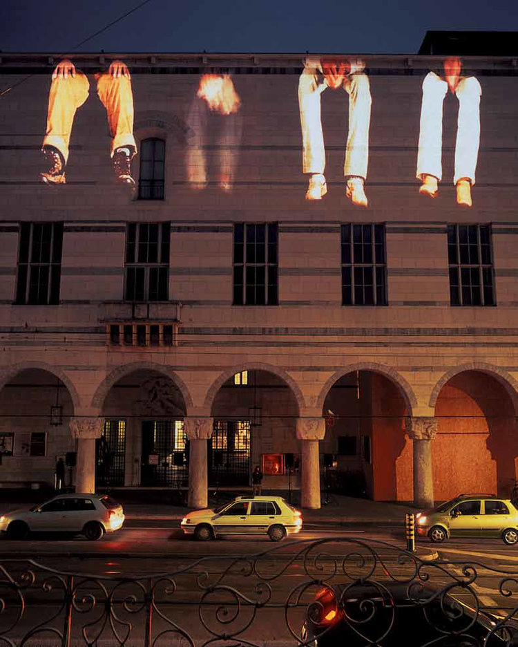 The facade of a building at night. Along the top half is projected the image of four people’s legs and feet, giving the appearance that they are sitting on the roof of the building with their legs hanging over the side.