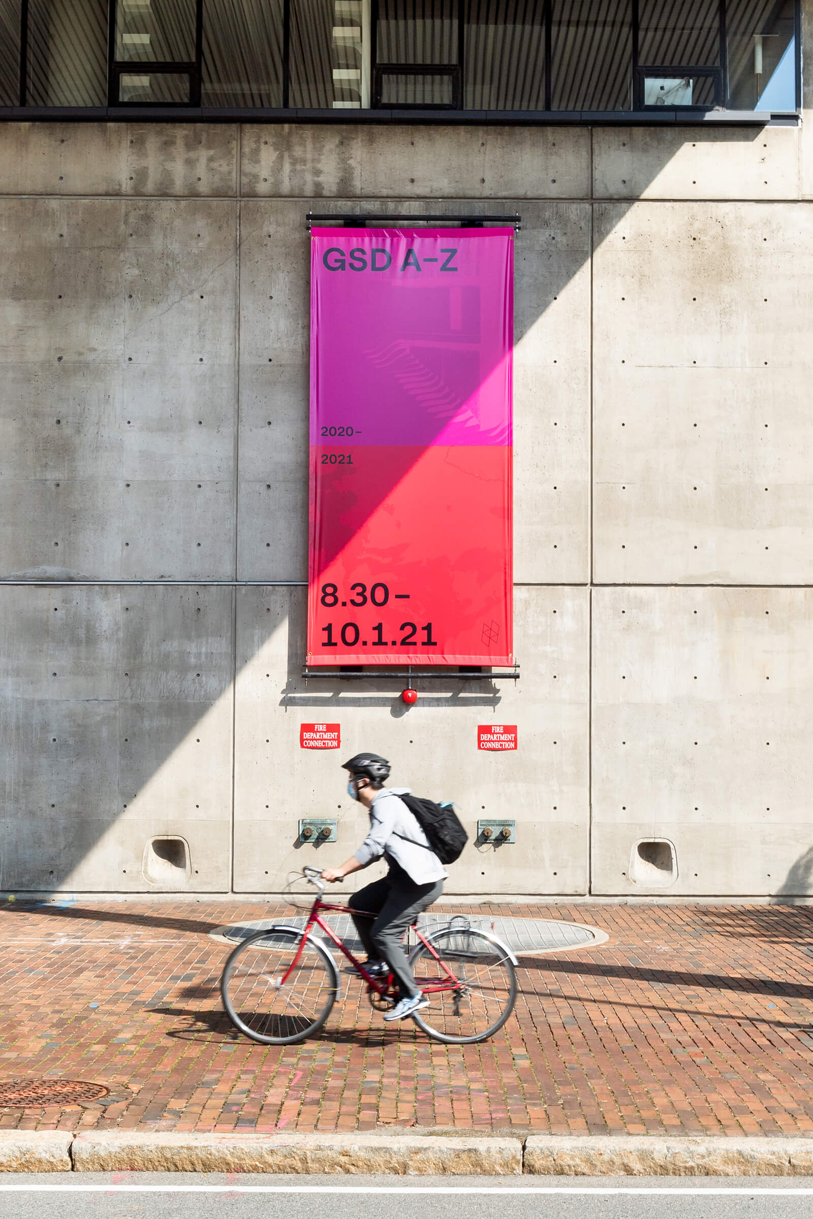 A student bikes past the A-Z exhibition poster hung on the outside of Gund Hall.