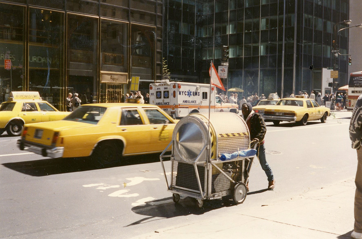 Photo of a busy street in New York City with yellow cabs driving by and people on the sidewalks. In the foreground, a person pushes one of Wodiczko’s Homeless Vehicles, a metal and wire cart that can carry belongings and unfold to create a sleeping area.